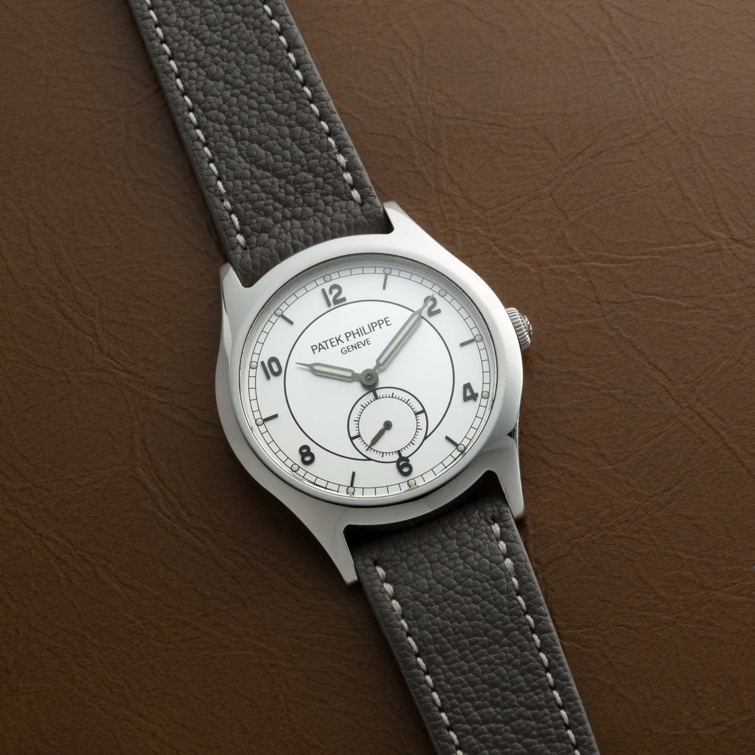 A Patek Philippe Calatrava Geneva Boutique Limited Edition wristwatch with a white dial and gray leather strap rests on a brown textured surface.
