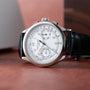 The Patek Philippe Chronograph, featuring a white dial, black leather strap, and silver case, is displayed on a brown surface.
