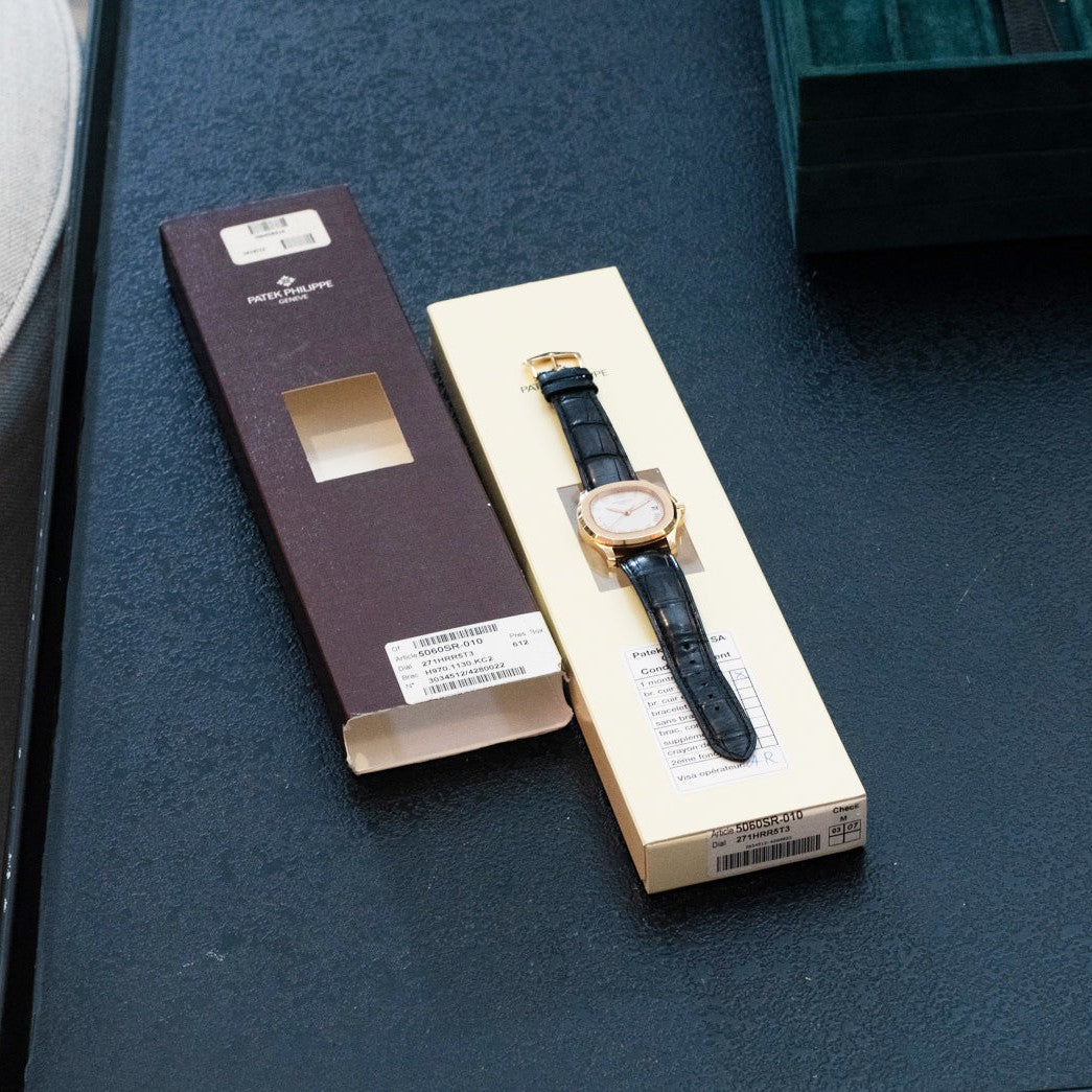 The Patek Philippe Aquanaut luxury wristwatch, in a cream box beside its brown case, rests on a dark table.