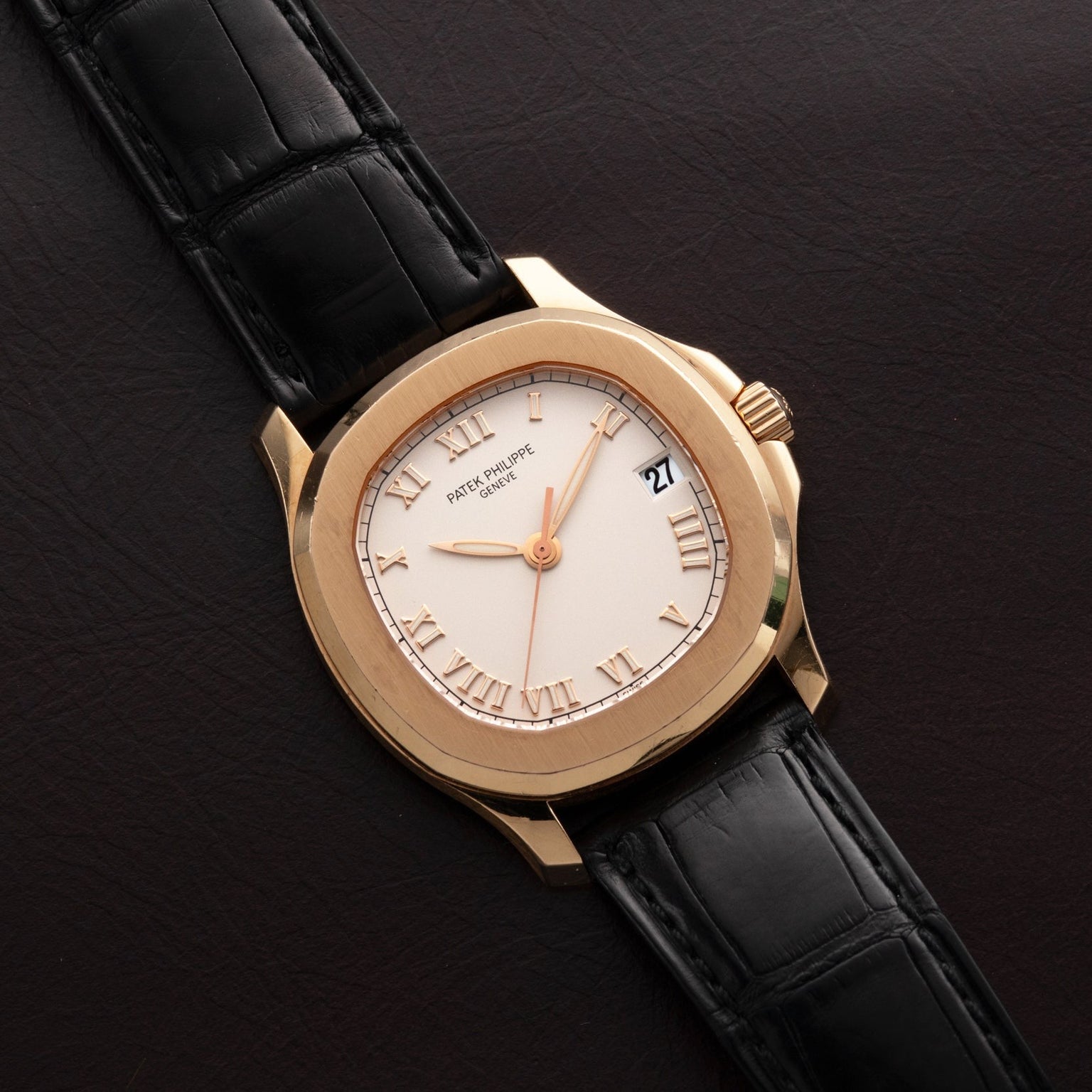 The Patek Philippe Aquanaut gold wristwatch with black leather strap rests on a dark brown leather surface, displaying Roman numerals and date.