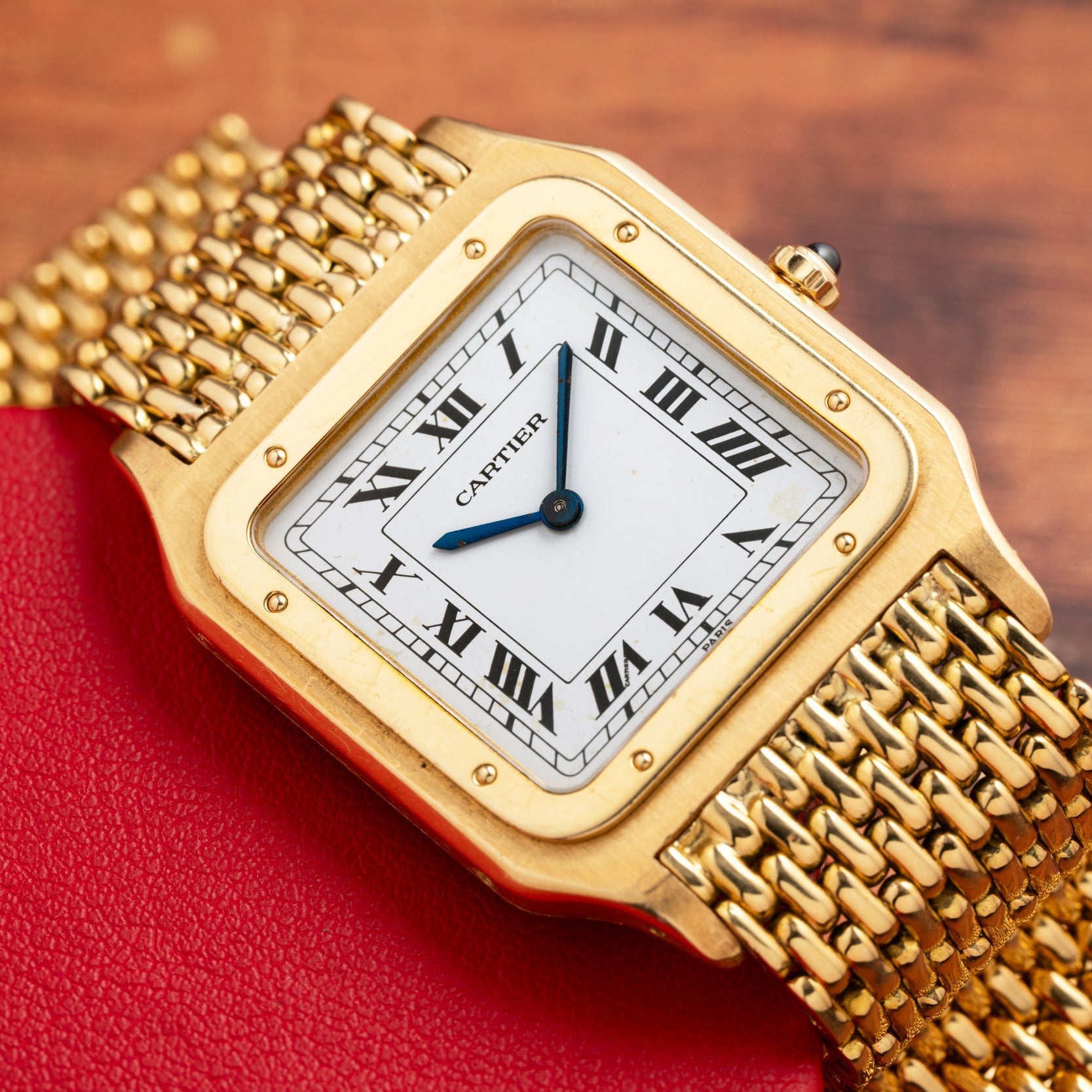 The Cartier Santos Dumont watch features a square face with Roman numerals and a woven metal band, shown resting on a red surface.