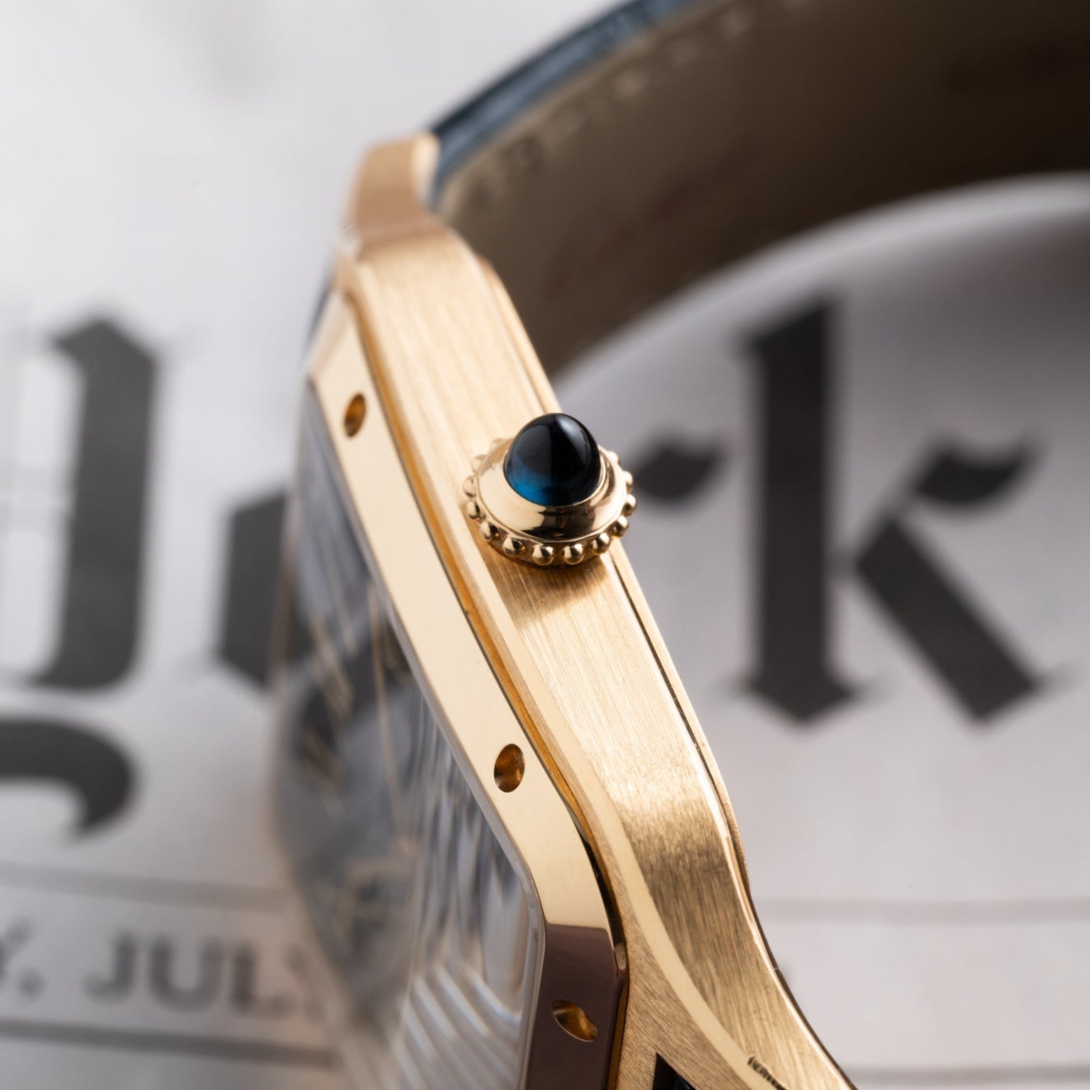 Close-up of the Cartier Santos Dumont gold wristwatch with a blue and gold crown, resting on a newspaper.