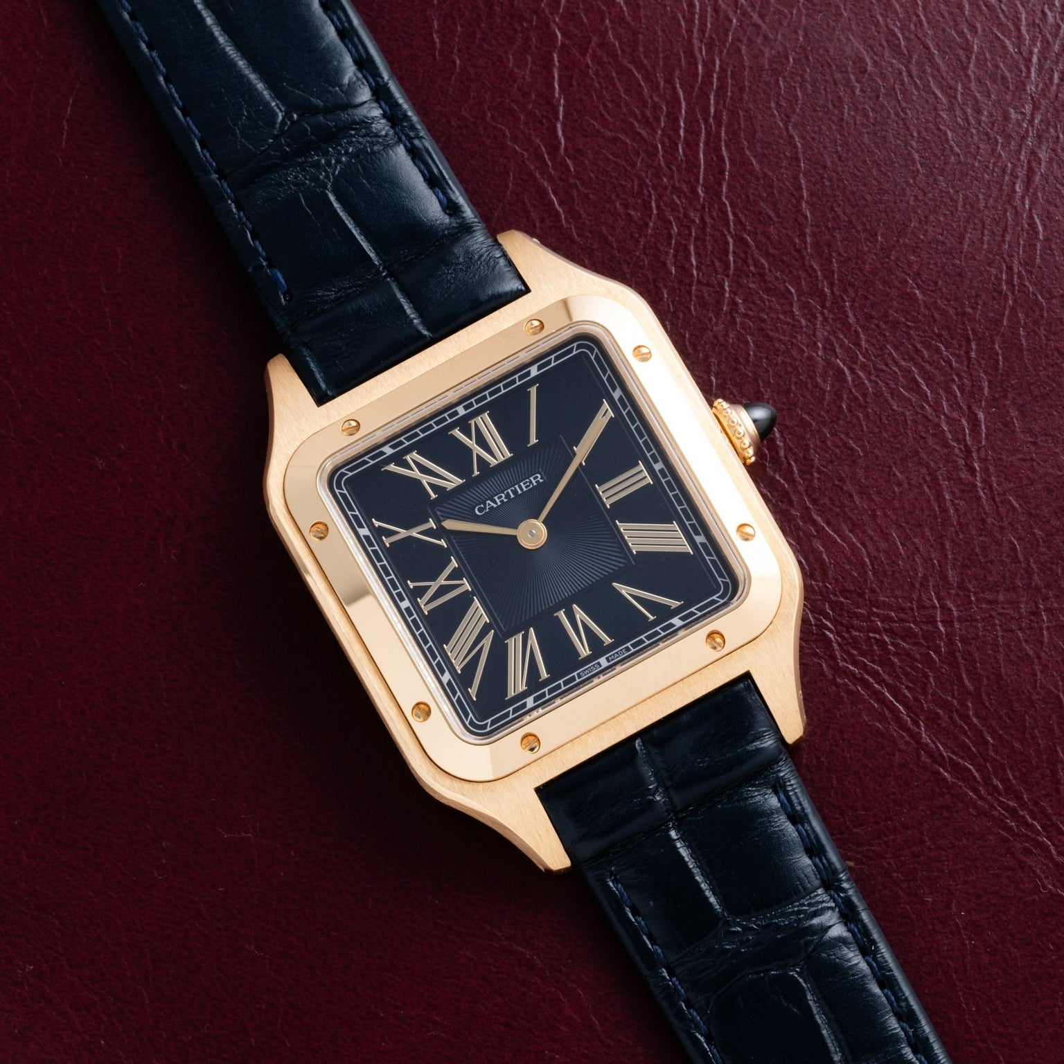The Cartier Santos Dumont watch, featuring a black leather strap and Roman numerals, rests on a dark red textured surface.