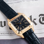 The Cartier Santos Dumont gold wristwatch with a black leather band rests on a newspaper displaying part of the word Wednesday.