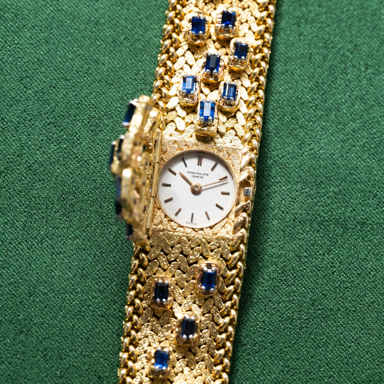 Patek Philippe Yellow Gold Integrated Dress Watch with blue gemstone accents on a green fabric background. The watch features a small, round face.