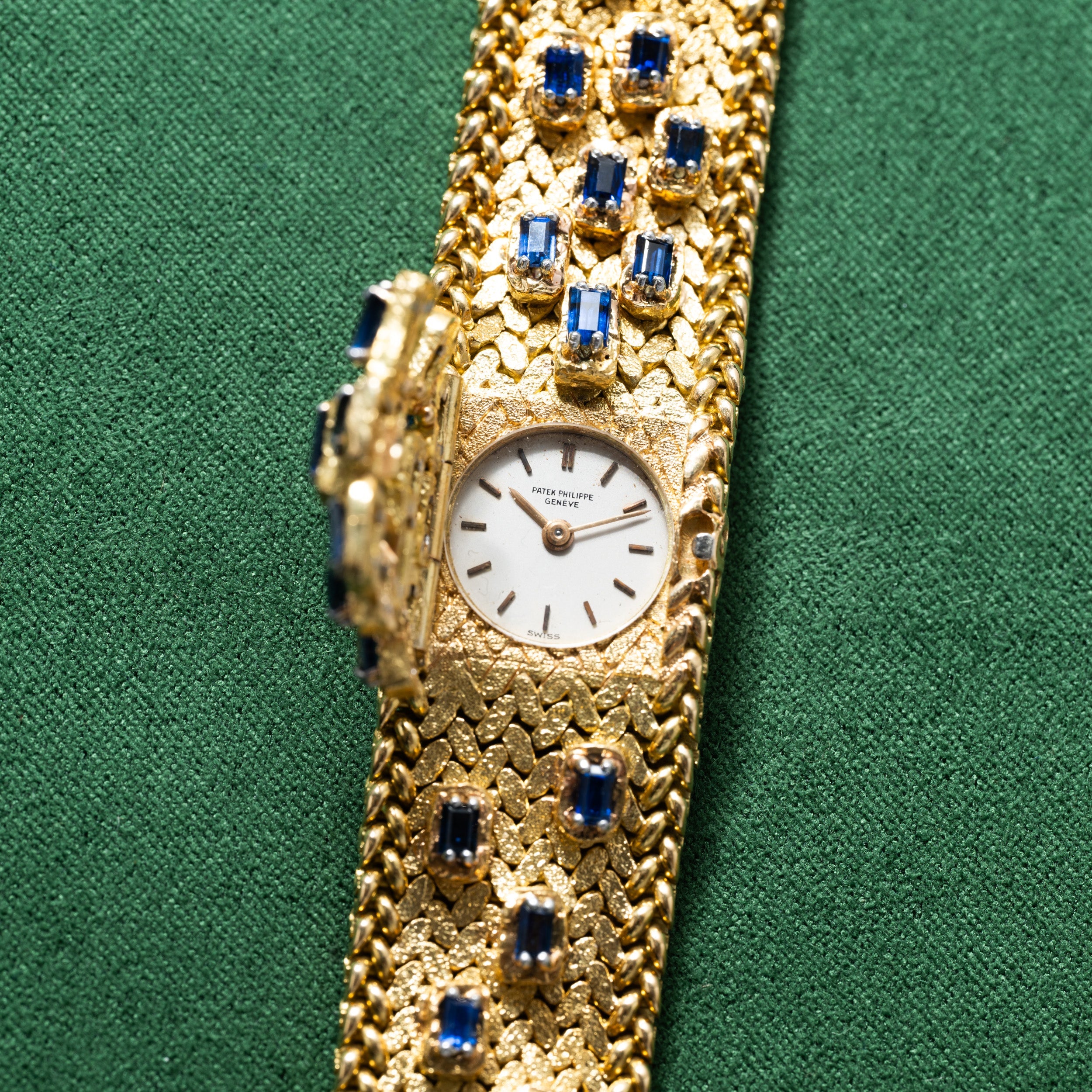 Patek Philippe Yellow Gold Integrated Dress Watch with blue gemstone accents on a green fabric background. The watch features a small, round face.