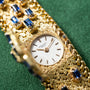A Patek Philippe Yellow Gold Integrated Dress Watch displayed on a textured green fabric background with blue gemstones.