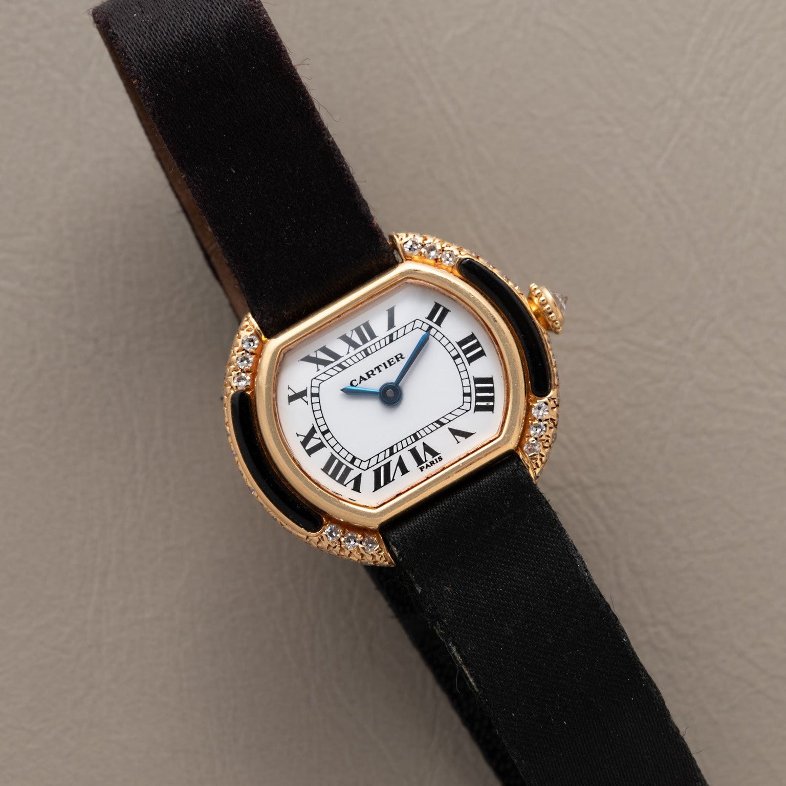 Cartier Ellipse watch featuring a black strap, gold case, Roman numerals, and diamond accents displayed on a beige surface.