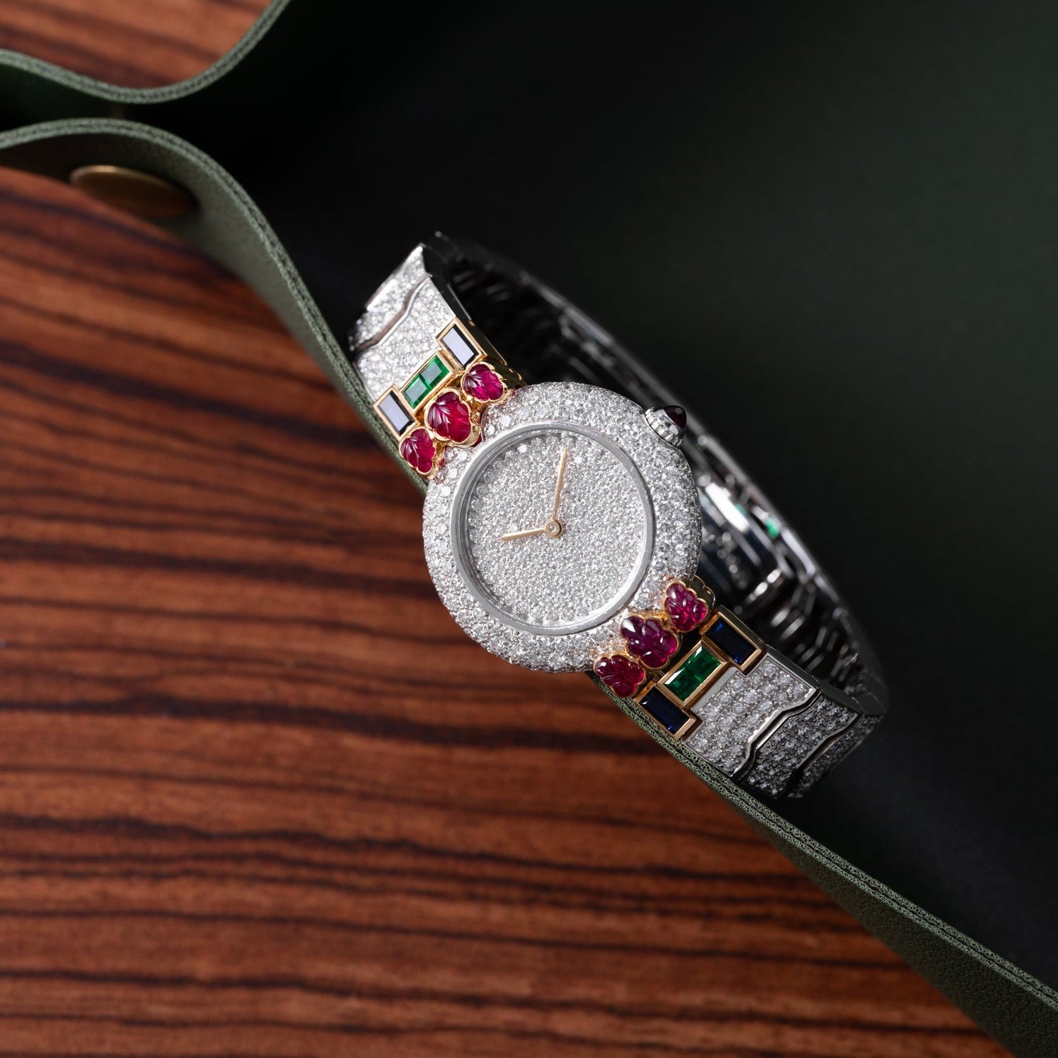A Cartier Colisee Tutti Frutti Diamond wristwatch adorned with diamonds, rubies, and emeralds rests on a wooden surface beside a green case.