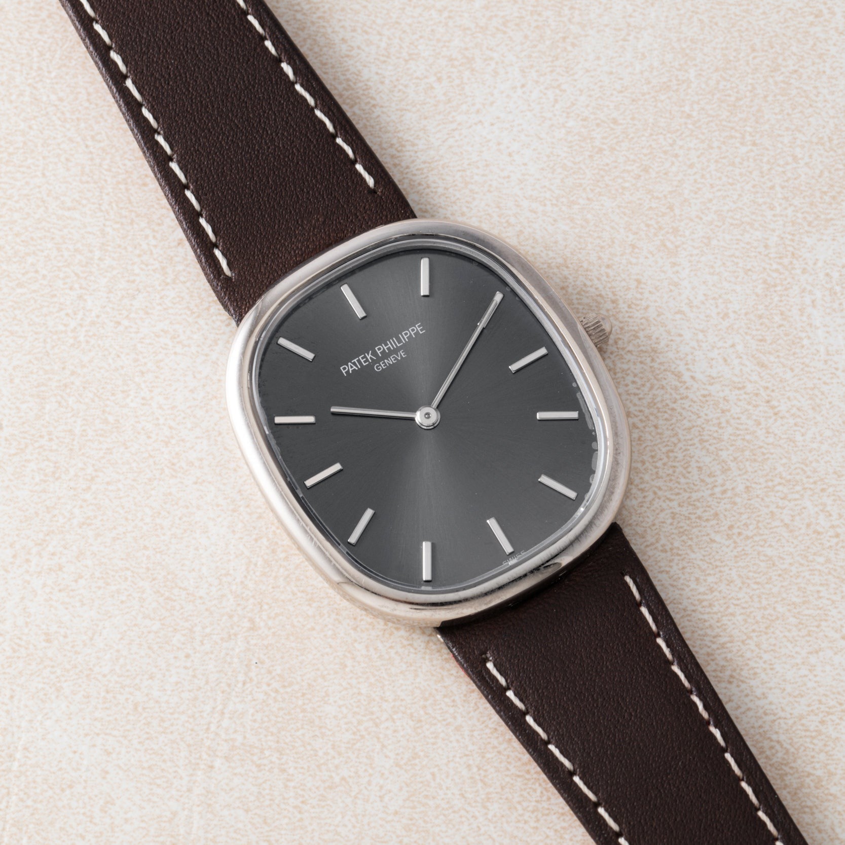 The Patek Philippe Ellipse watch features a dark dial and a brown leather strap, displayed on a light textured surface.