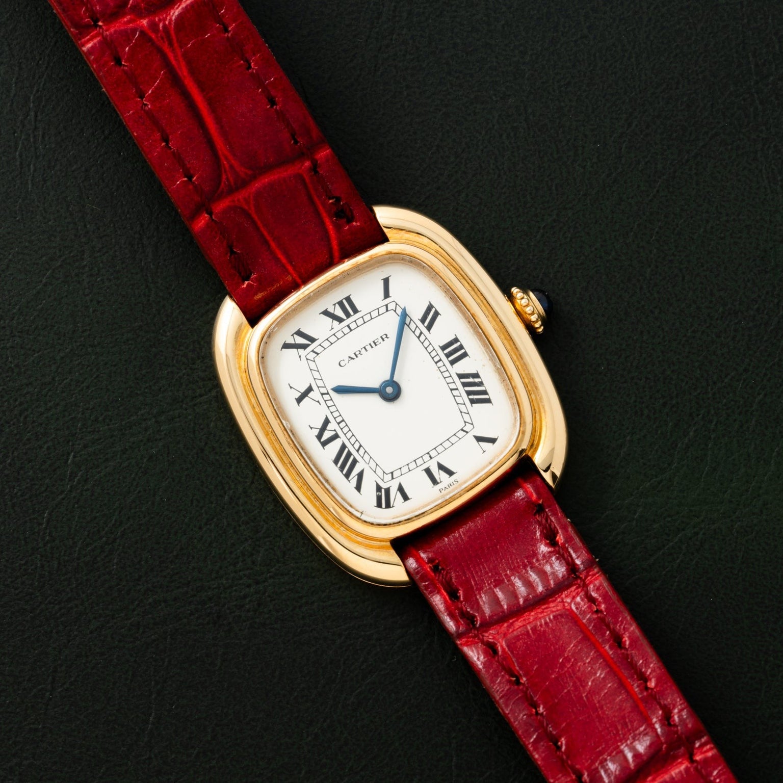 A Cartier Gondole Ladies gold watch with a red leather strap rests on a dark green surface.