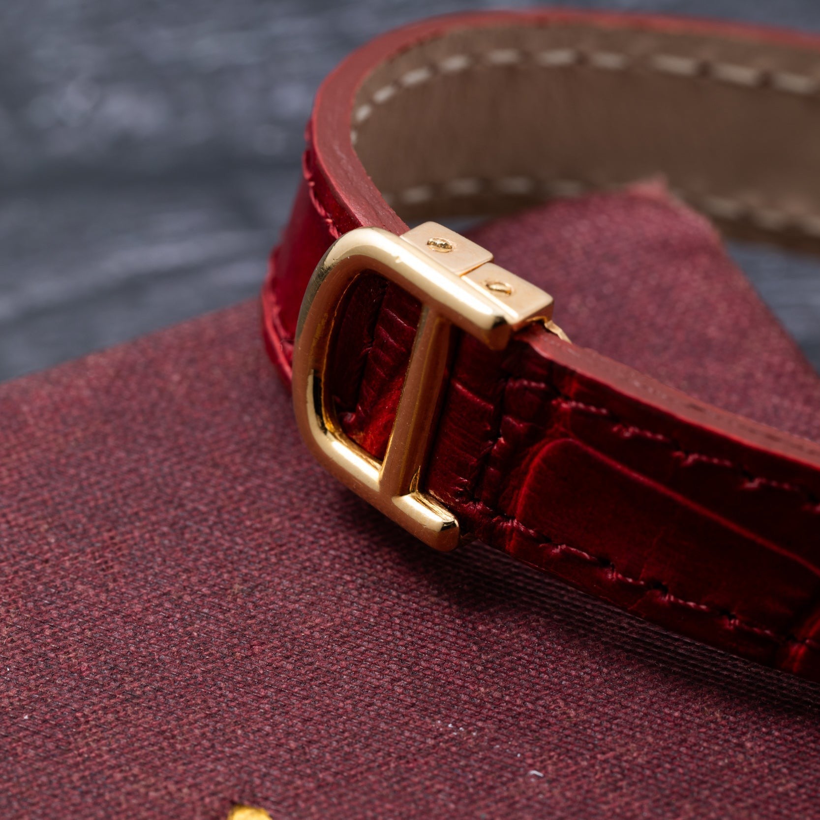 Close-up of the gold buckle on a Cartier Gondole Ladies red leather watch strap, resting on a textured maroon surface.