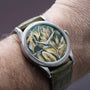 The Patek Philippe Calatrava Cloisonné Tropical Leaves II is a wristwatch with a green and gold leaf-patterned dial on a green leather strap, shown worn on a person’s wrist.