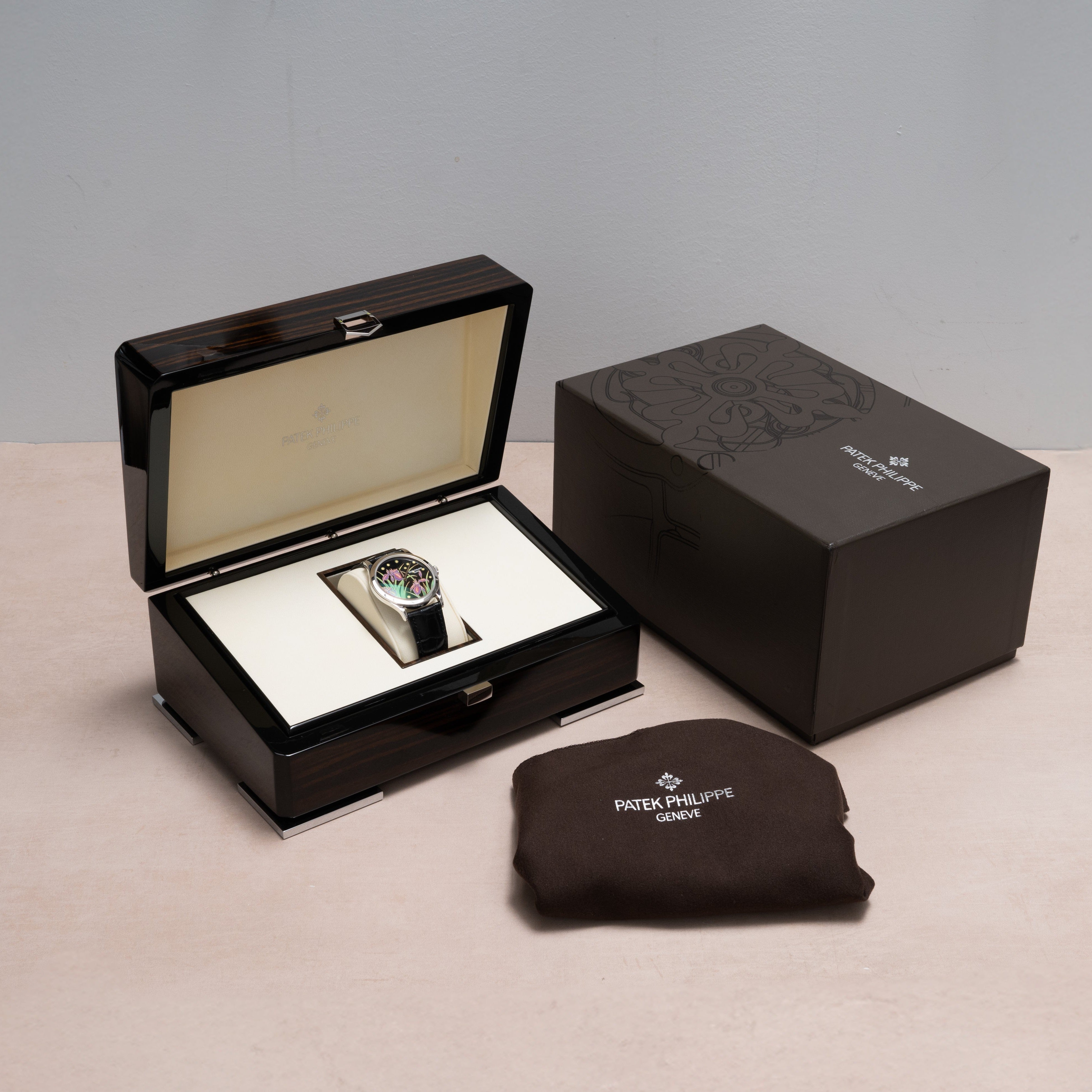 A Patek Philippe Calatrava Cloisonné Iris & Night Fireflies watch is displayed in its box, with the outer box and branded cloth placed nearby on a beige surface.