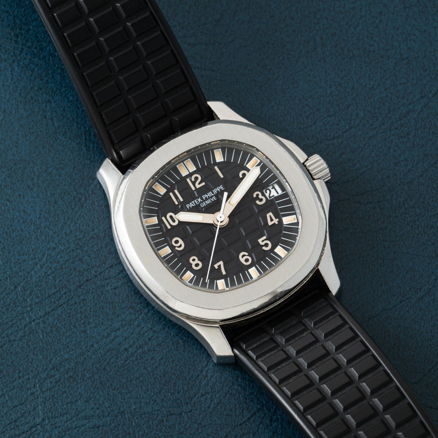 A Patek Philippe Aquanaut watch featuring a black dial and rubber strap displayed on a textured dark blue surface.
