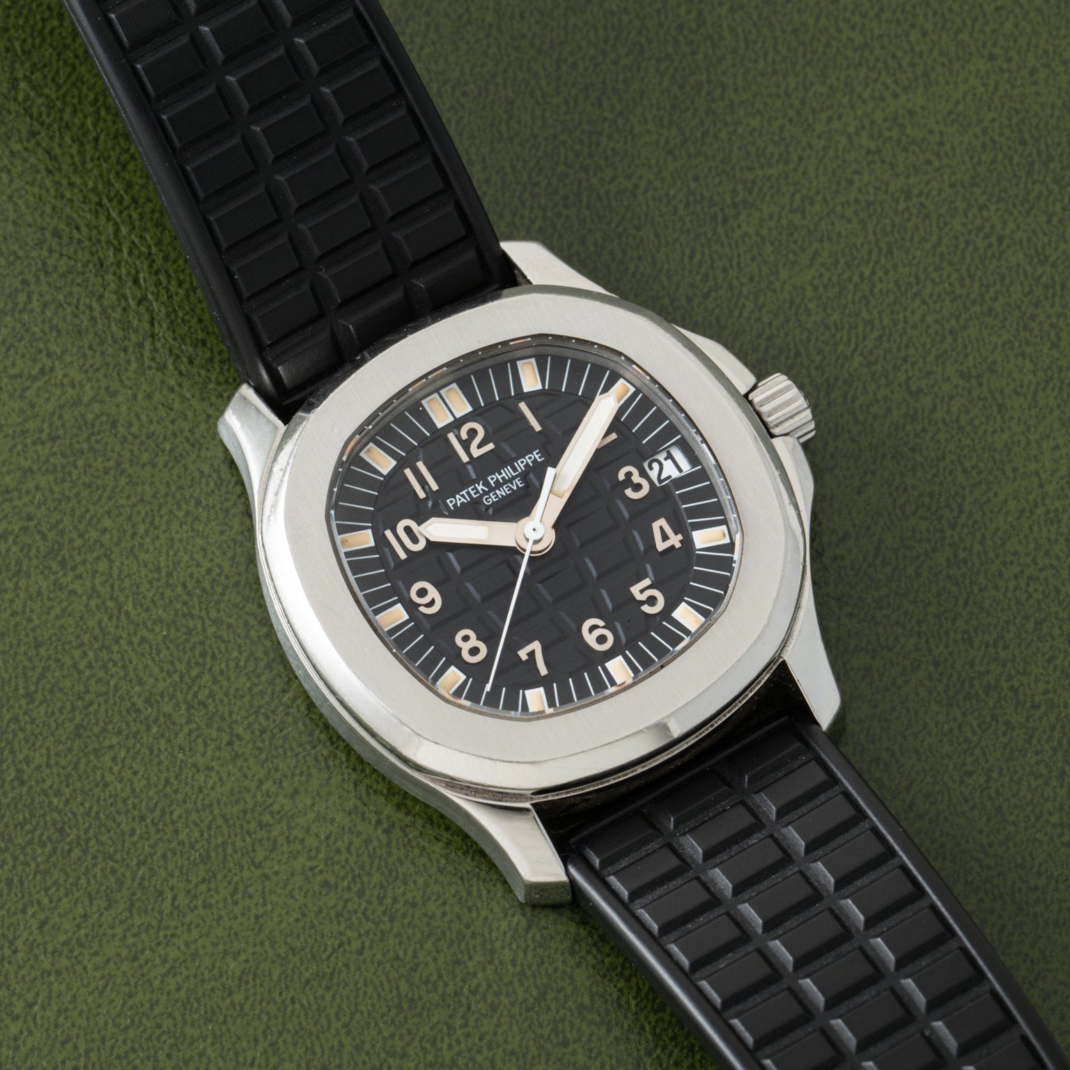 A black Patek Philippe Aquanaut wristwatch with a rubber strap rests on a textured green surface.