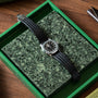 The Patek Philippe Aquanaut, a black wristwatch with a grid-pattern strap, is displayed on a green speckled surface inside a green box.