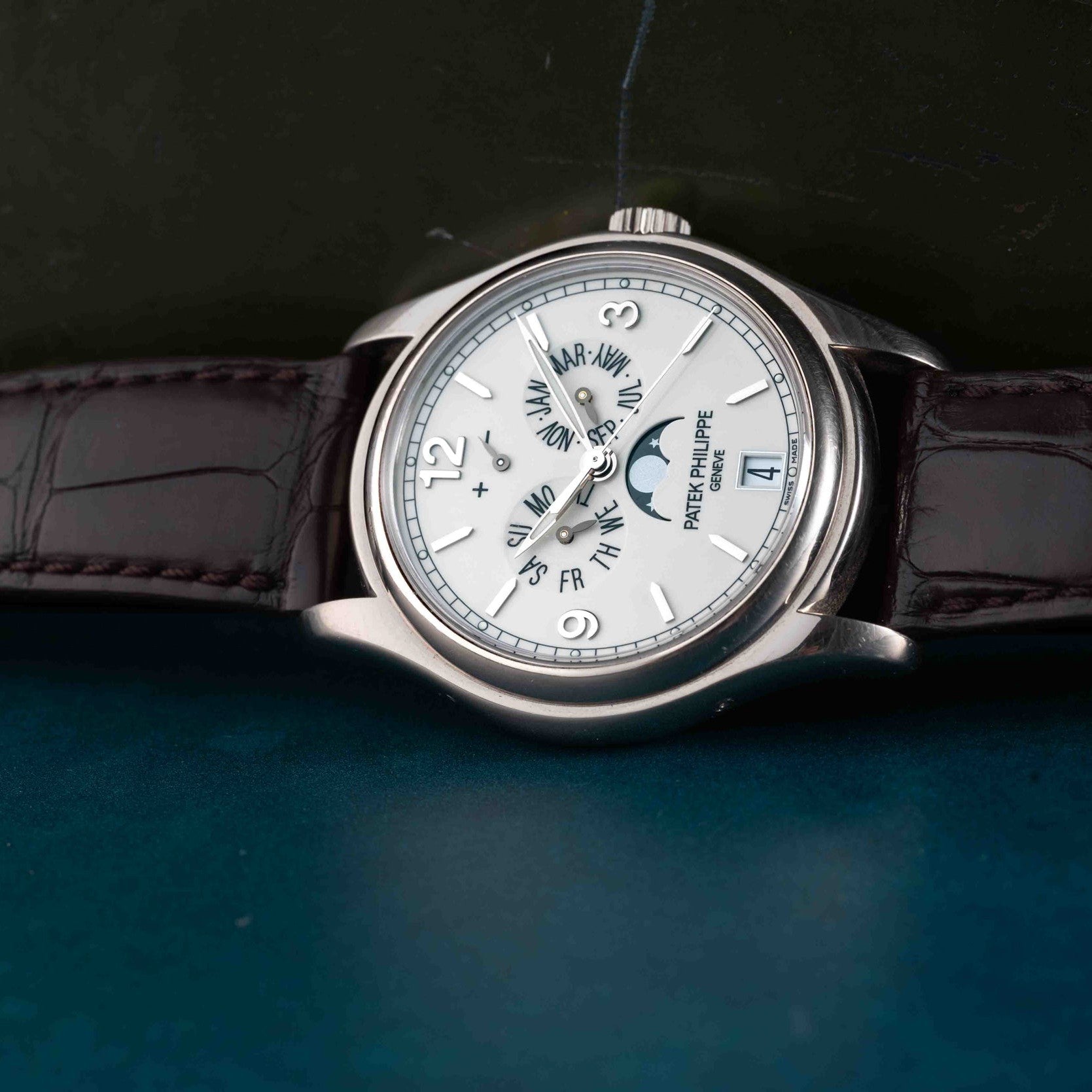 The Patek Philippe Annual Calendar Moonphase features a silver case, white dial, moon phase display, and a dark leather strap, displayed on a blue surface.