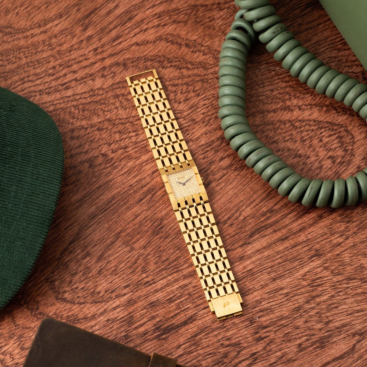 A Piaget Dancer gold wristwatch with a linked band rests on wood beside green items and a coiled phone cord.