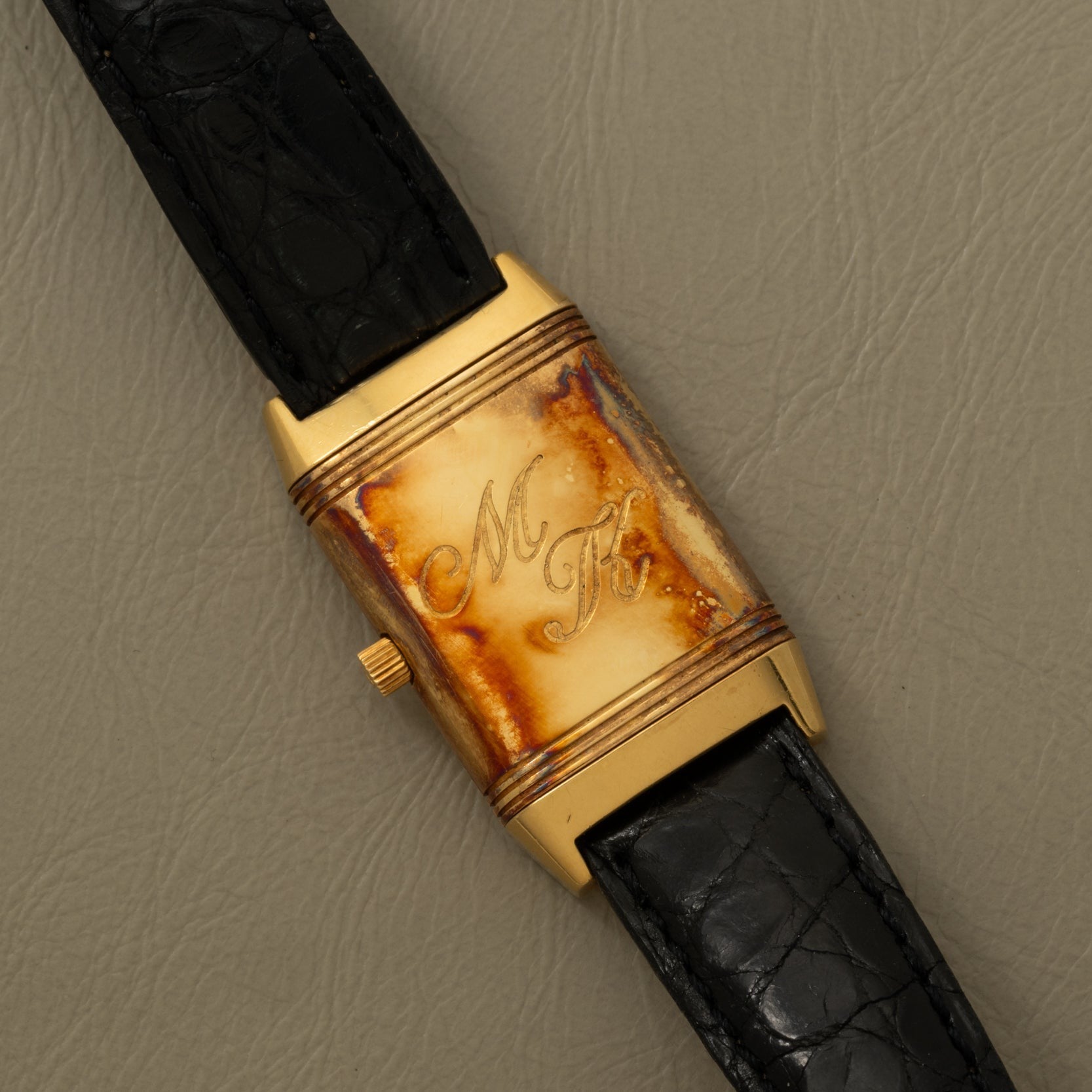 A Jaeger-LeCoultre Reverso Classique gold watch with a rectangular engraved “M T K” face and a black leather strap is displayed on a gray surface.