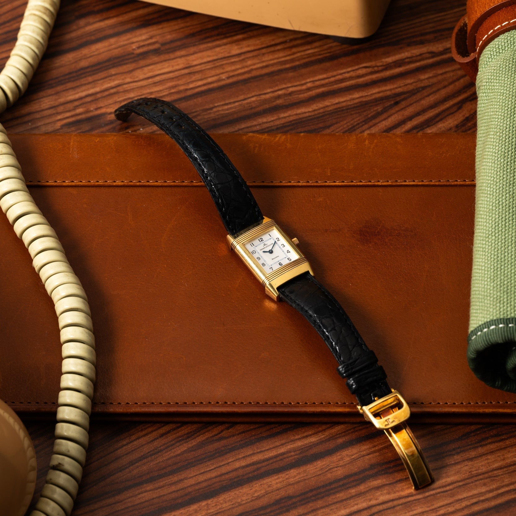 A Jaeger-LeCoultre Reverso Classique gold wristwatch with a black leather strap rests on a brown leather mat, next to a phone cord and green fabric.