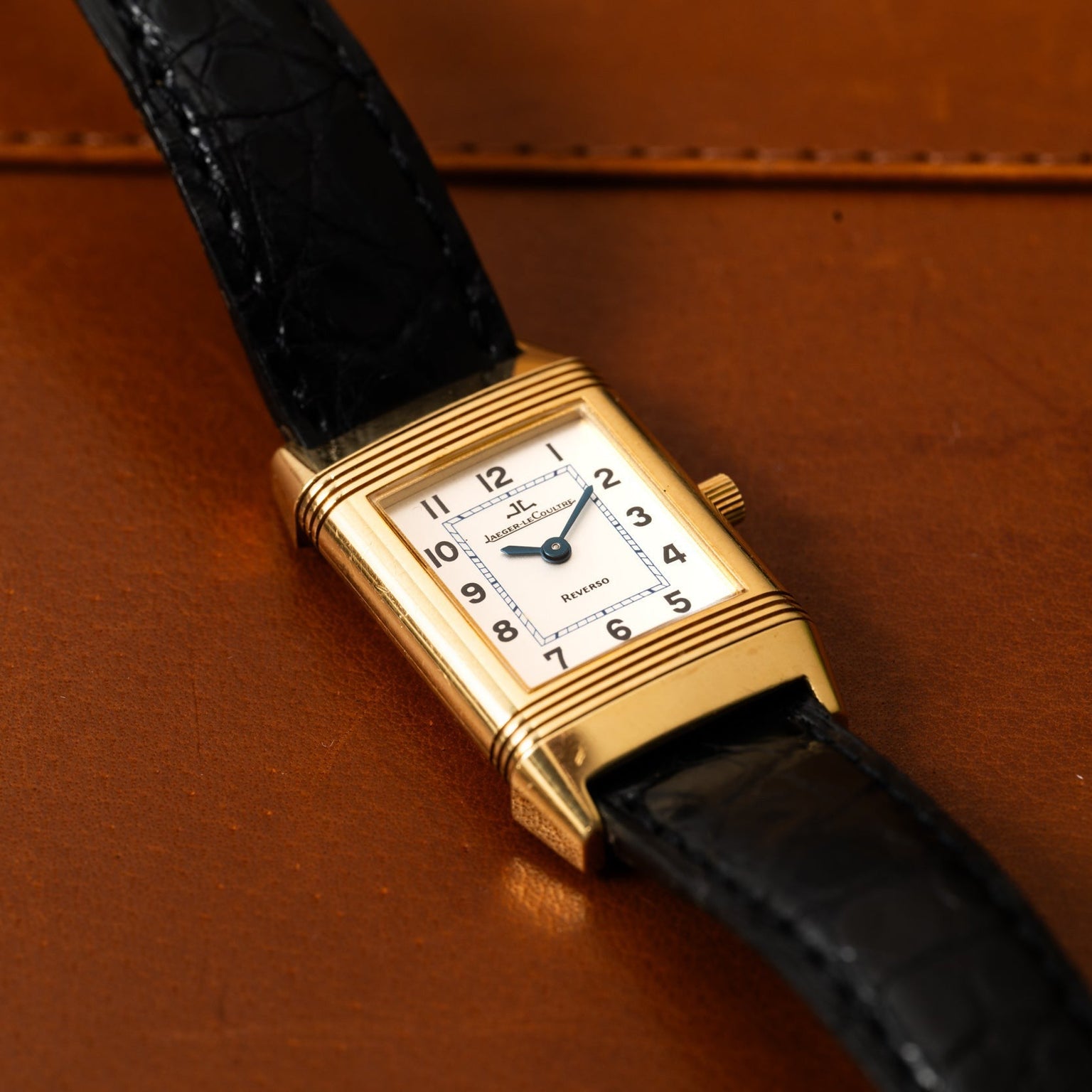 A Jaeger-LeCoultre Reverso Classique wristwatch with a gold rectangular case, white dial, and black leather strap displayed on a brown leather surface.