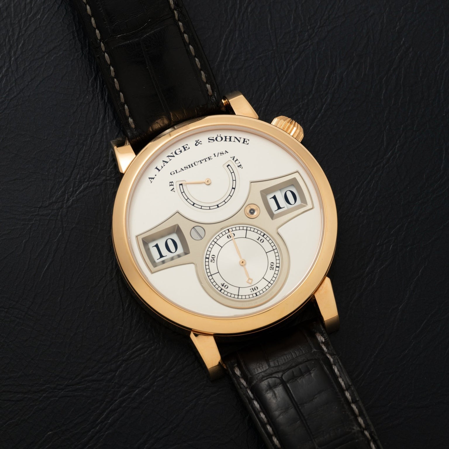 The Lange & Sohne Zeitwerk watch features a digital time display and black leather strap, shown on a black surface.