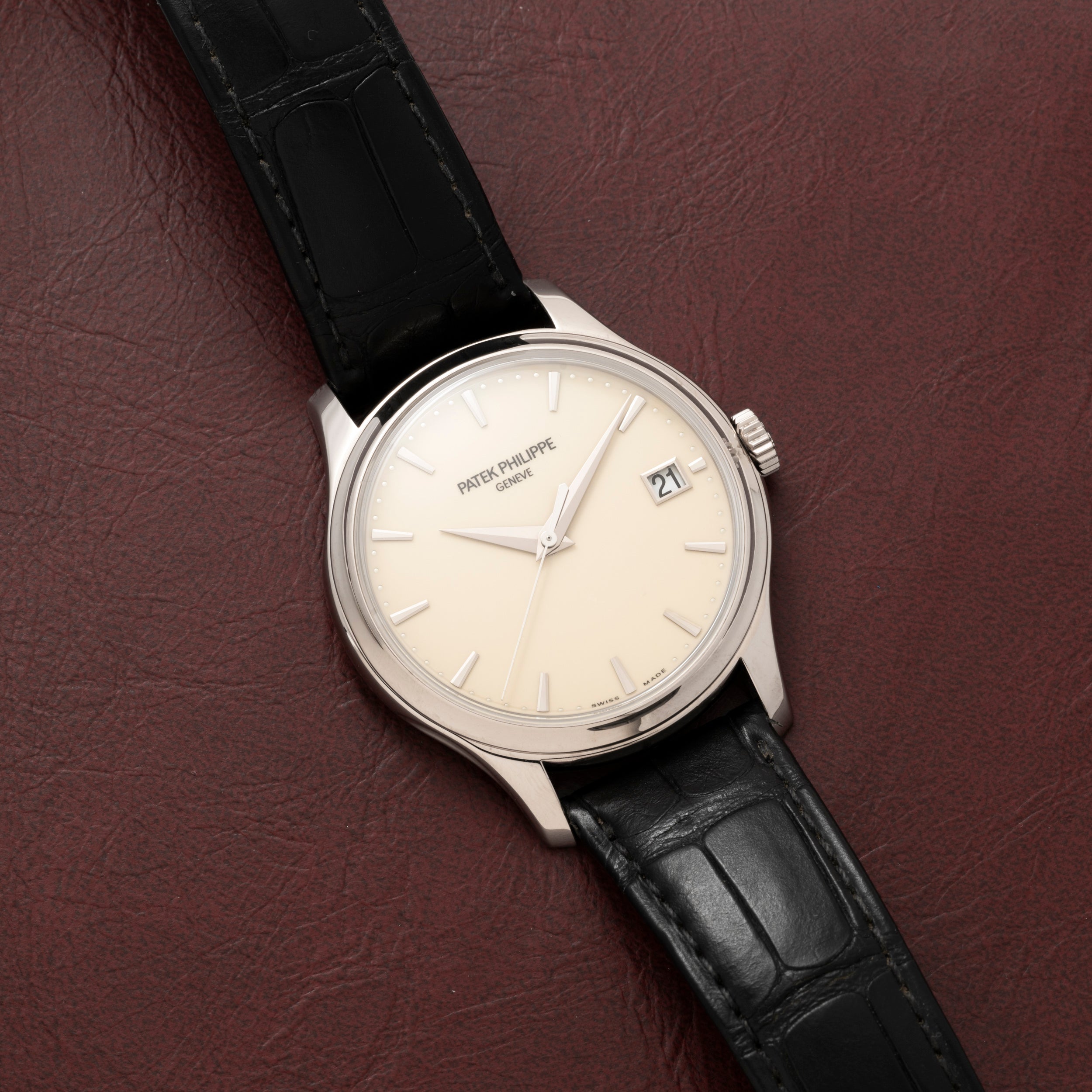 A Patek Philippe Calatrava wristwatch with a black leather strap rests on a brown leather surface, displaying the date 27.