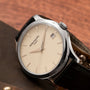 A Patek Philippe Calatrava wristwatch with a cream dial, date display, and black leather strap rests on a brown surface.