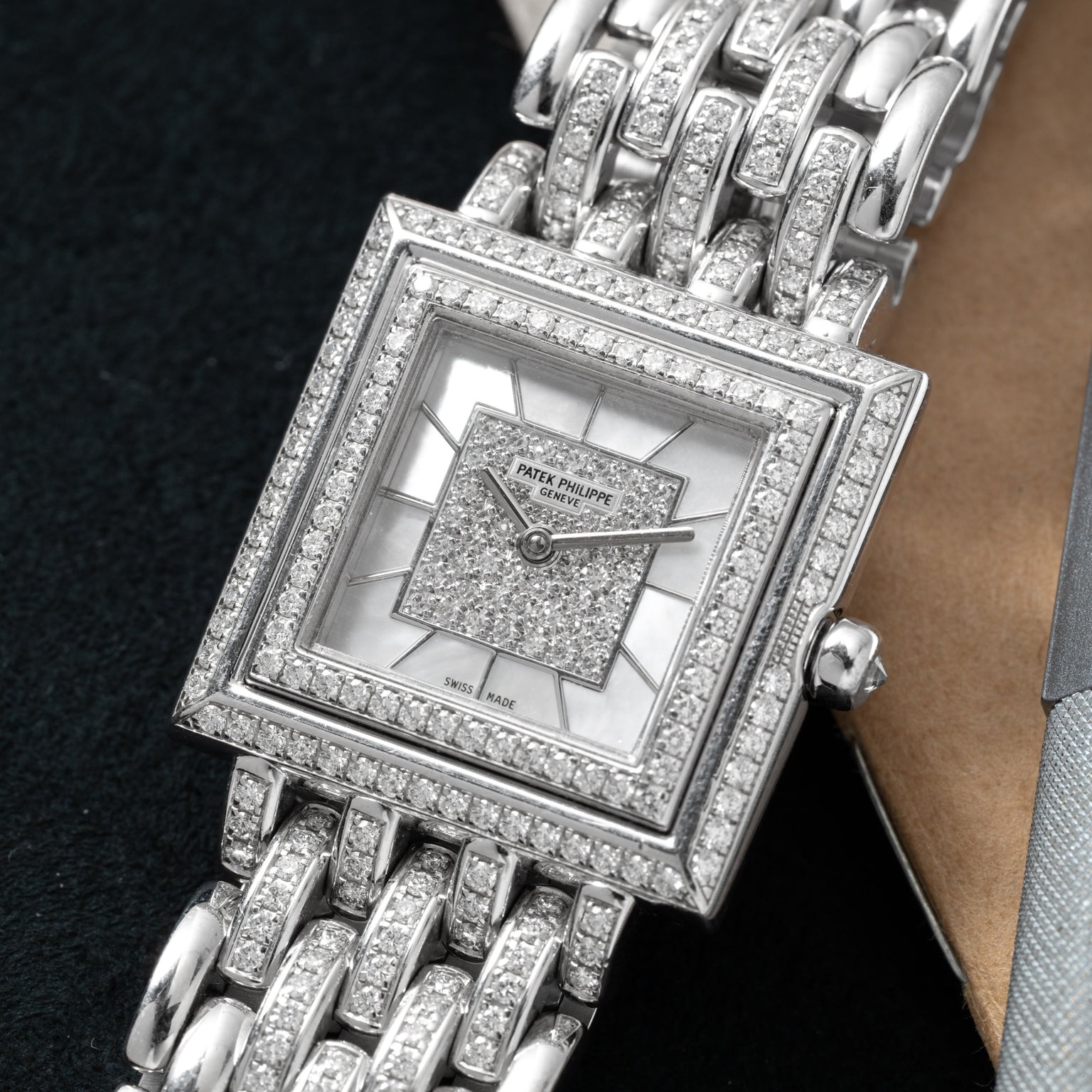 A square Patek Philippe Gondolo watch with diamond accents and a metal link band rests on a dark surface.