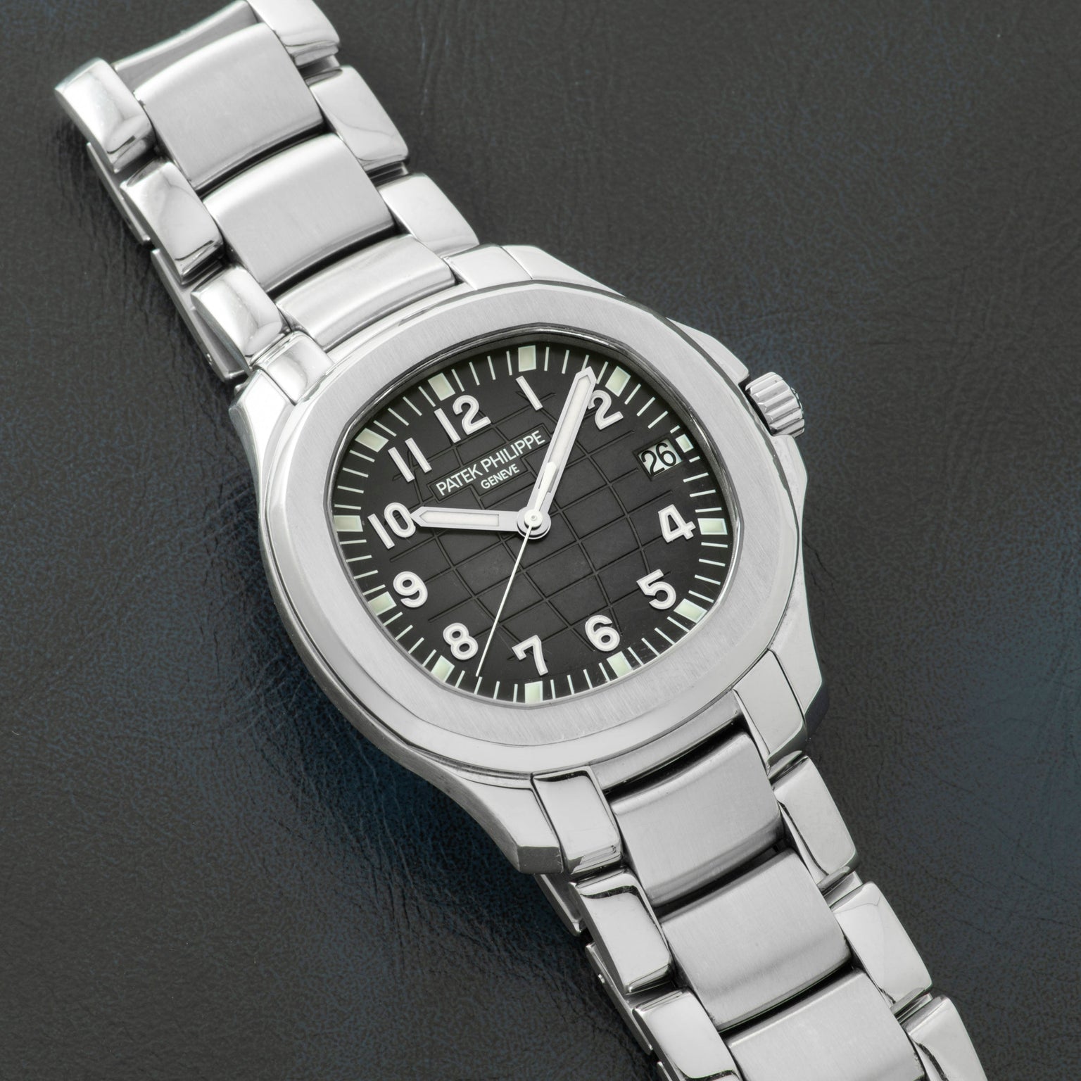A Patek Philippe Aquanaut stainless steel wristwatch with a black dial and metal bracelet displayed on a dark surface.