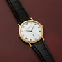 Patek Philippe Calatrava gold watch with black leather strap and Roman numerals displayed on a burgundy leather surface.