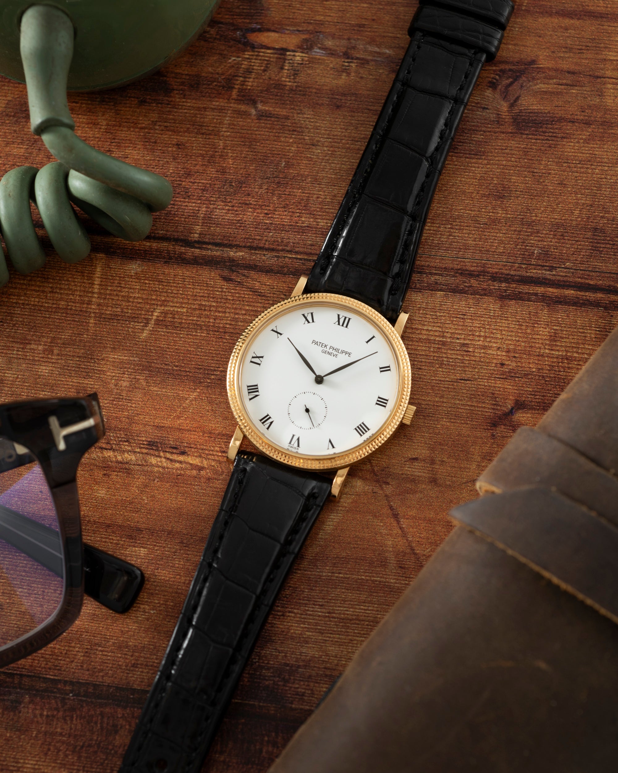 A Patek Philippe Calatrava gold watch with a black leather strap rests on a wooden surface beside glasses and a coiled green cord.