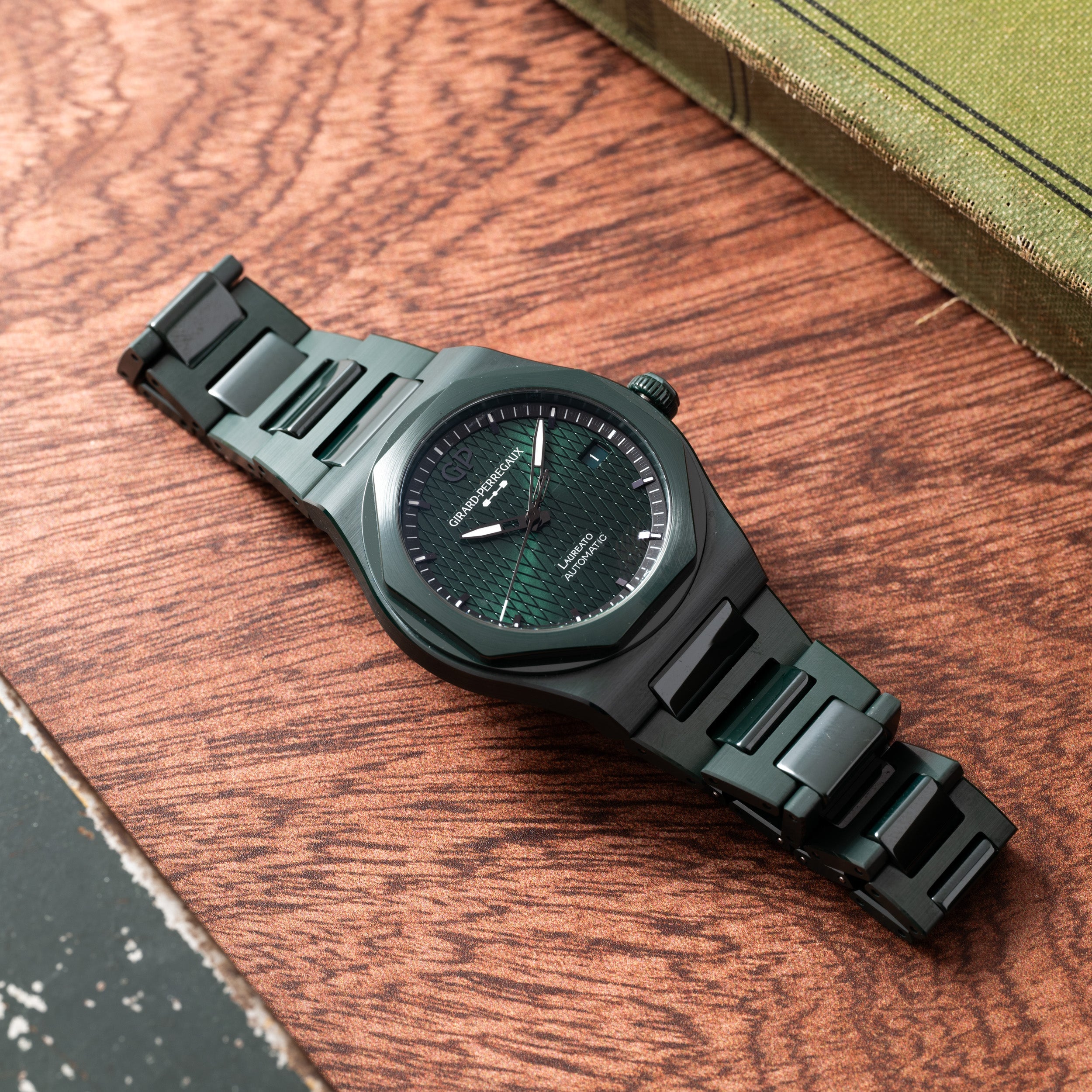 The Laureato Green Ceramic Aston Martin Edition by Girard-Perregaux, with its textured green dial, rests on a wooden surface near a green book.