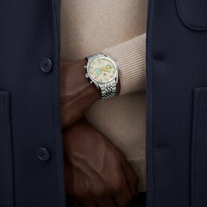 Silver stainless steel chronograph watch with cream dial and subdials, worn on wrist between navy and beige jacket layers.