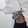 A person in a white shirt and jeans wears the TAG Heuer Limited Edition Carrera Skipper For HODINKEE with a blue face, date window, and gray fabric strap—a unique pick for collectors who love vintage chronographs.