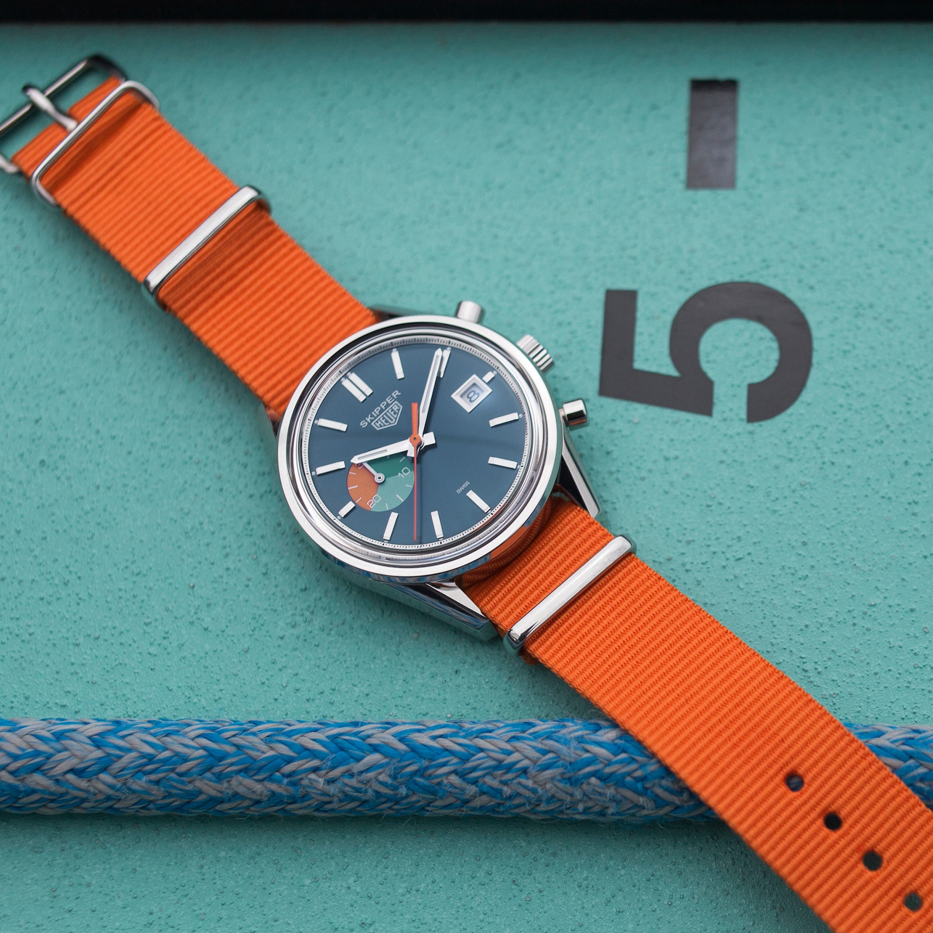 The TAG Heuer Limited Edition Carrera Skipper For HODINKEE, featuring an orange fabric strap and black dial, sits on a teal surface near a large 5 and blue-and-white braided rope—a bold pick for vintage chronograph enthusiasts.