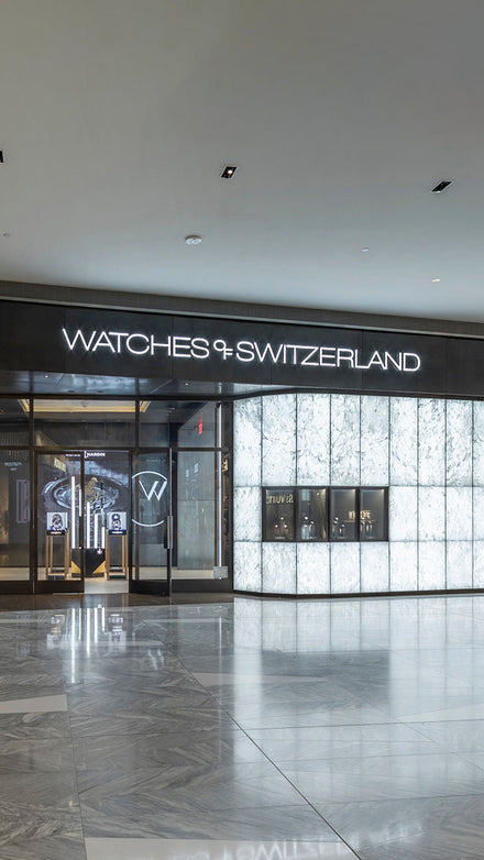 Storefront of Watches of Switzerland with illuminated signage and marble-patterned exterior walls.