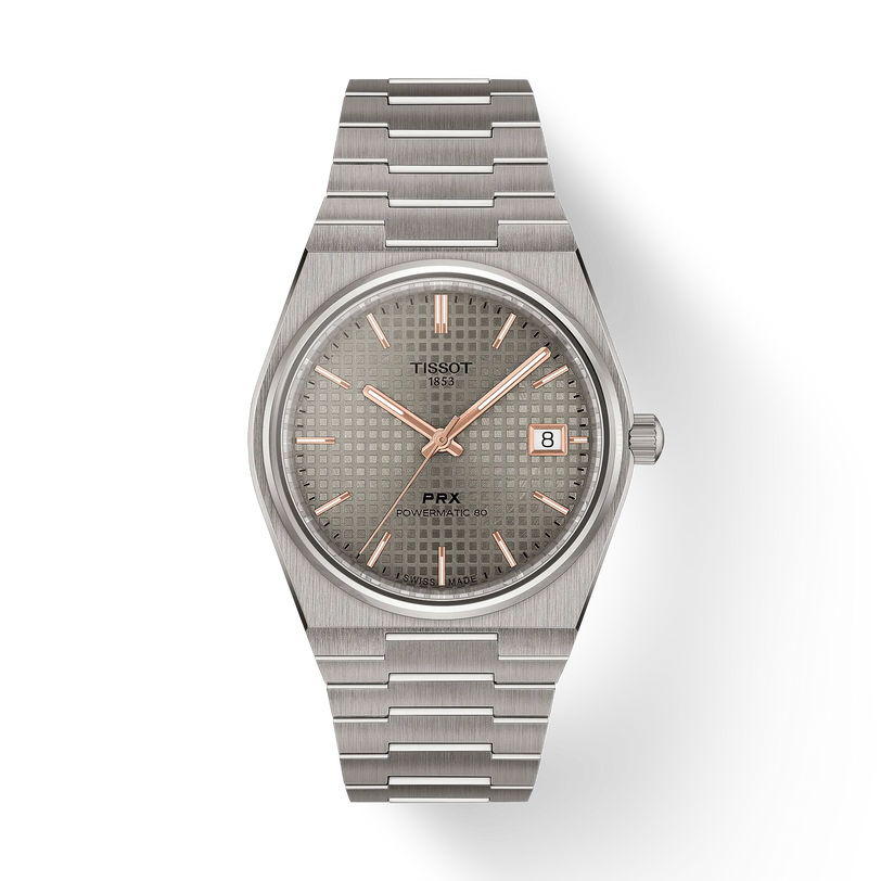 The Tissot PRX 38 features a stainless steel bracelet, silver textured dial, gold markers and hands, sapphire crystal, and a date window at 3 o’clock. The watch is showcased against a plain white background.