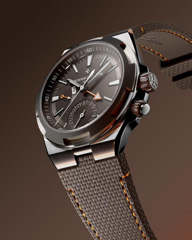 Steel watch with dark dial, dual time subdial, orange accents, and gray fabric strap with orange stitching.