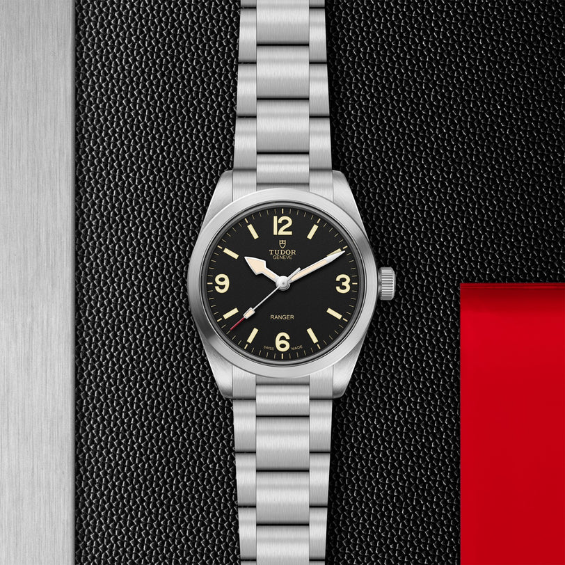 A TUDOR Ranger stainless steel watch with a black dial, luminous hour markers, and brushed metal bracelet is displayed on a textured black surface featuring a red accent on the right.