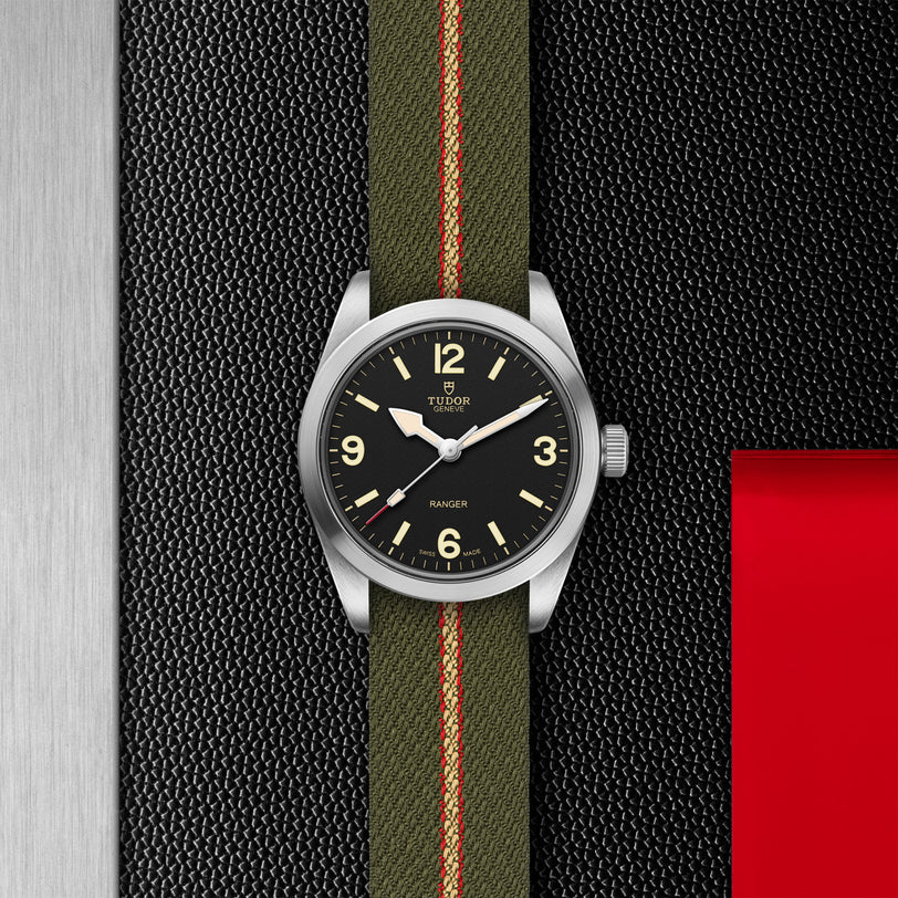 The TUDOR Ranger watch features a stainless steel case, black dial with large cream numerals, and an olive green fabric strap with red and beige stripes, shown on a textured black and silver background with a red accent.