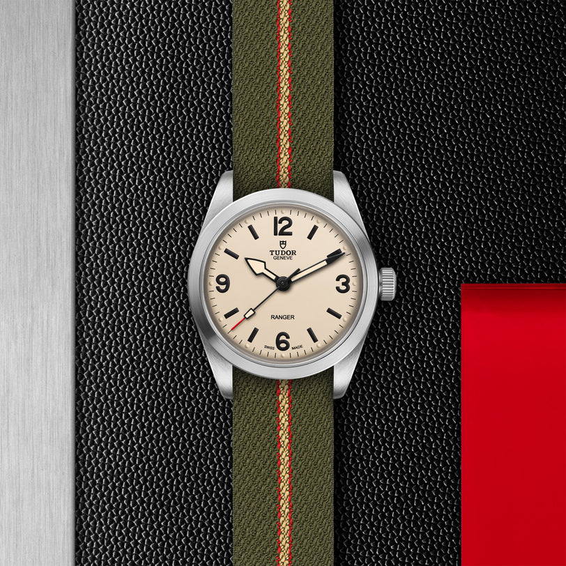 The TUDOR Ranger expedition watch features a cream dial with black numerals and a green fabric strap with red and beige stripes, set against textured black, red, and metallic backgrounds that highlight its robust stainless steel case.
