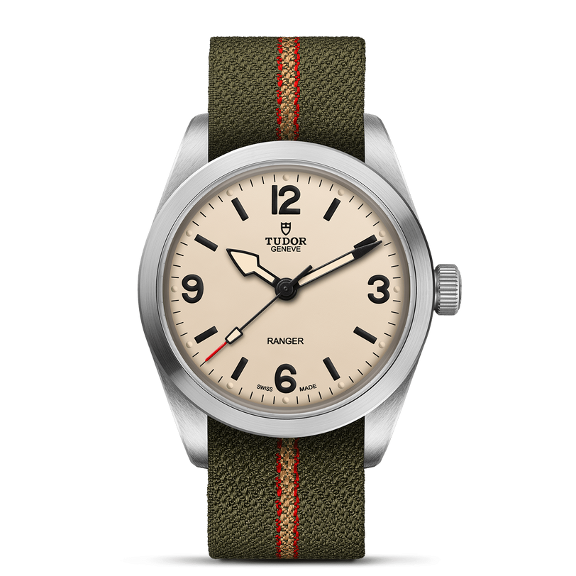 The TUDOR Ranger is an expedition watch with a beige dial, bold black numerals and hands, a stainless steel case, and an olive-green fabric strap with a red center stripe.
