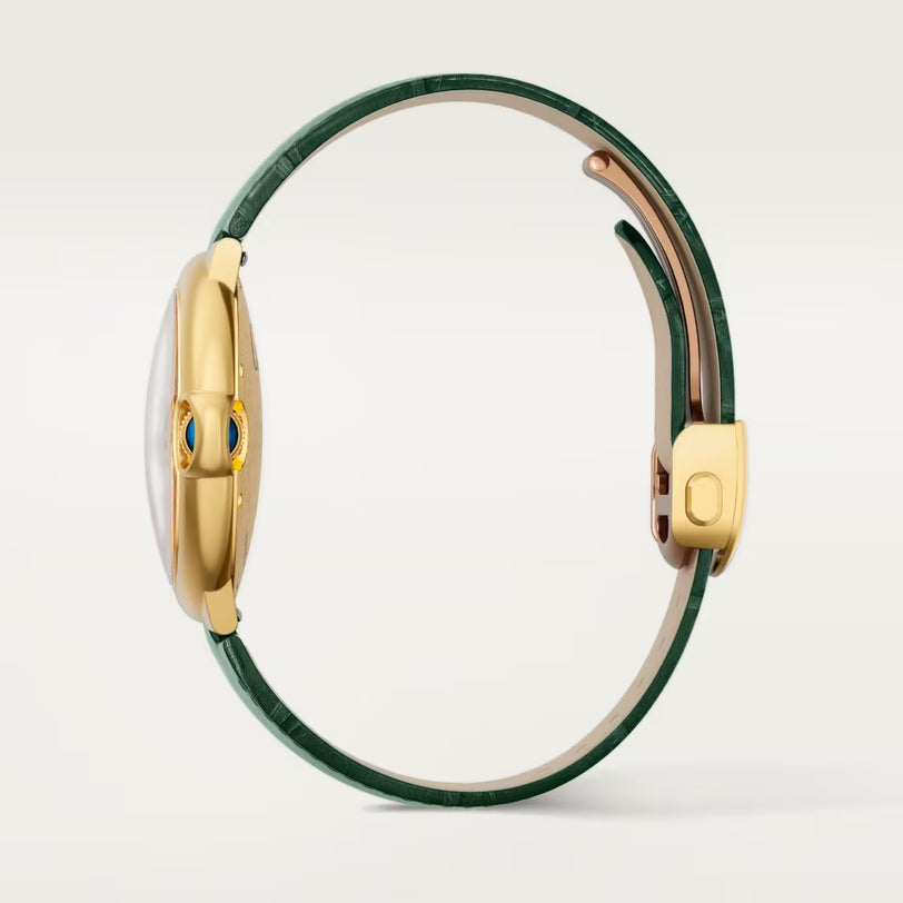 A side view of the Cartier Ballon Bleu De Cartier Watch featuring a gold case, dark green leather strap, and blue gem accents on the crown, set against a light background.