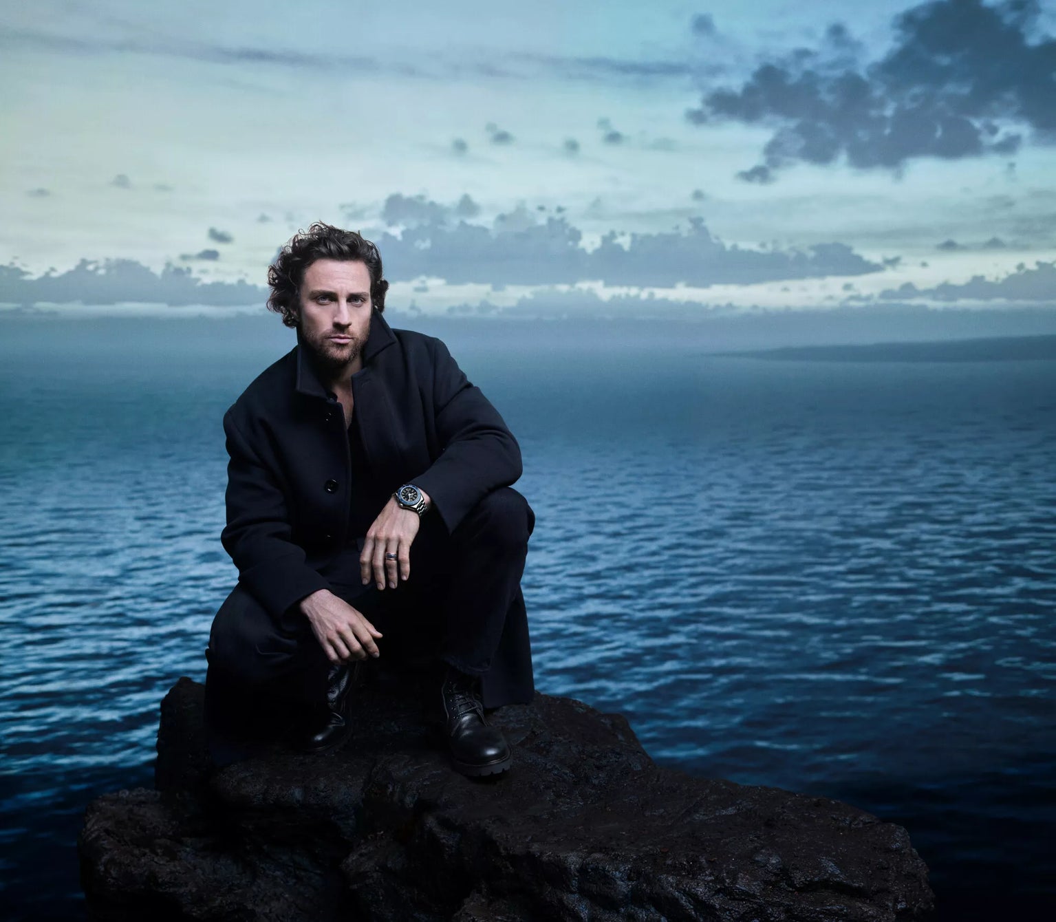 A bearded man with curly hair, dressed in dark clothing and boots, sits on a rocky cliff at twilight, his OMEGA Seamaster Planet Ocean 600mm watch catching the last light as he gazes over the calm ocean under a cloudy sky.
