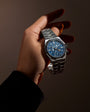 Stainless steel watch with blue dial, dual time subdials, and metal bracelet held in hand.