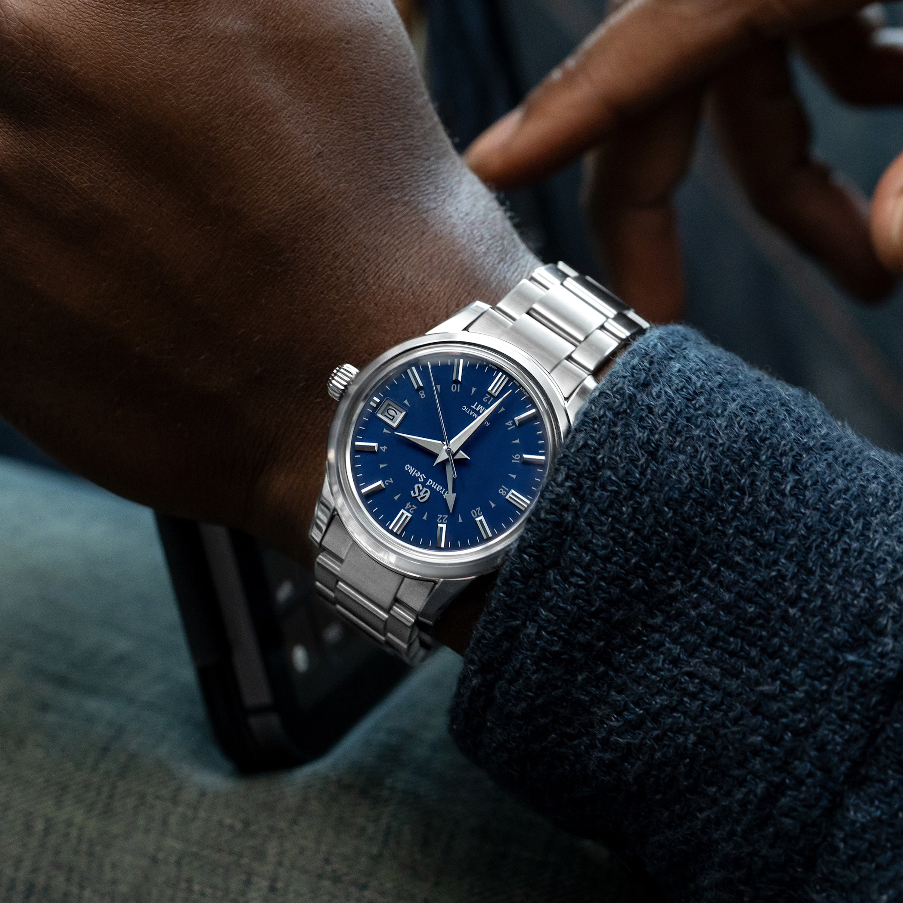 A close-up of a wrist wearing the Grand Seiko Automatic GMT SBGM239 Limited Edition For HODINKEE, featuring a blue dial and silver markers. The wearer, in a dark blue sweater, holds a black smartphone—an ideal look for Grand Seiko enthusiasts.