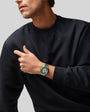 Man wearing black crewneck sweatshirt with green and silver sports watch on wrist.