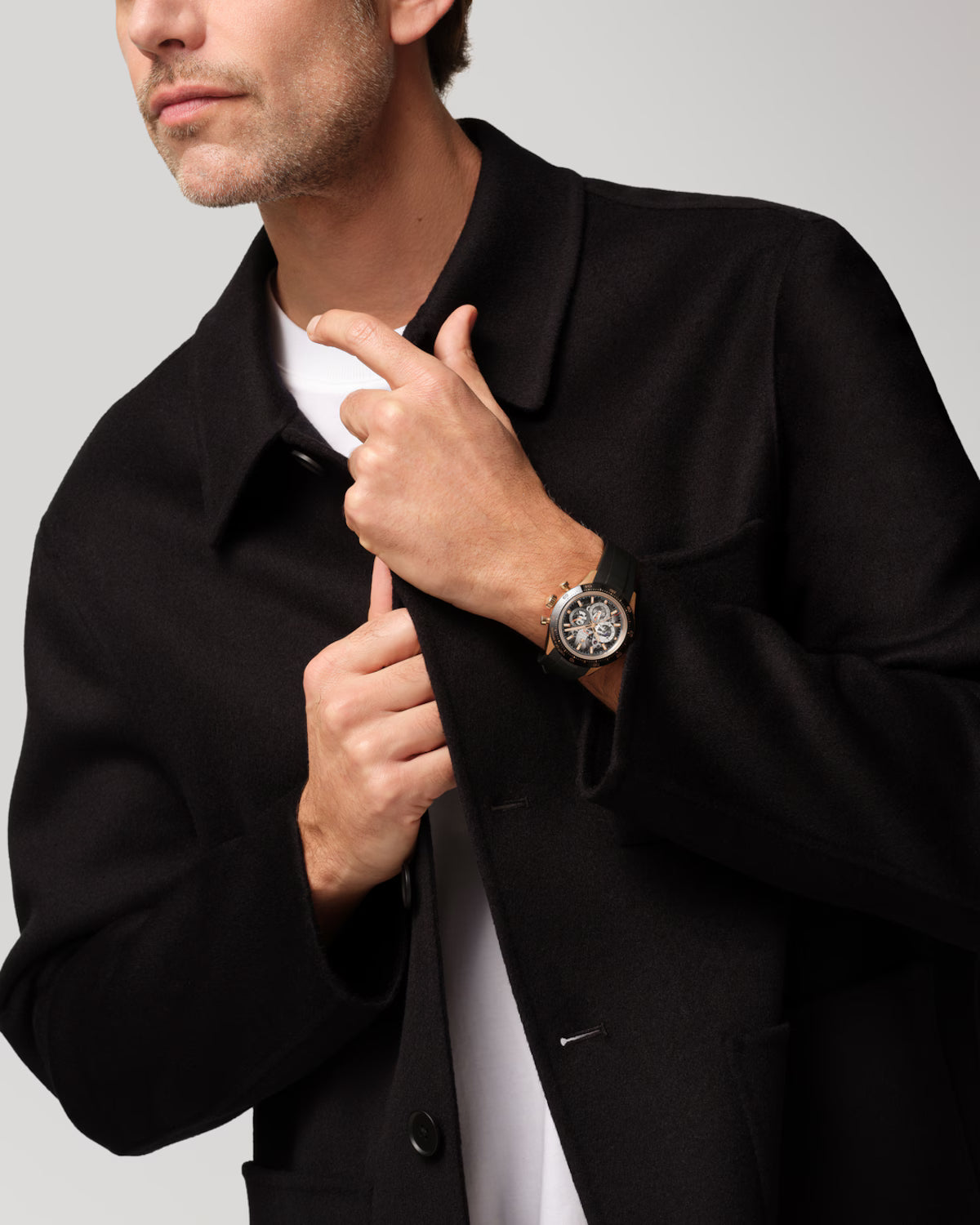 Man wearing black blazer with white shirt, displaying watch on wrist with white dial and brown leather band.